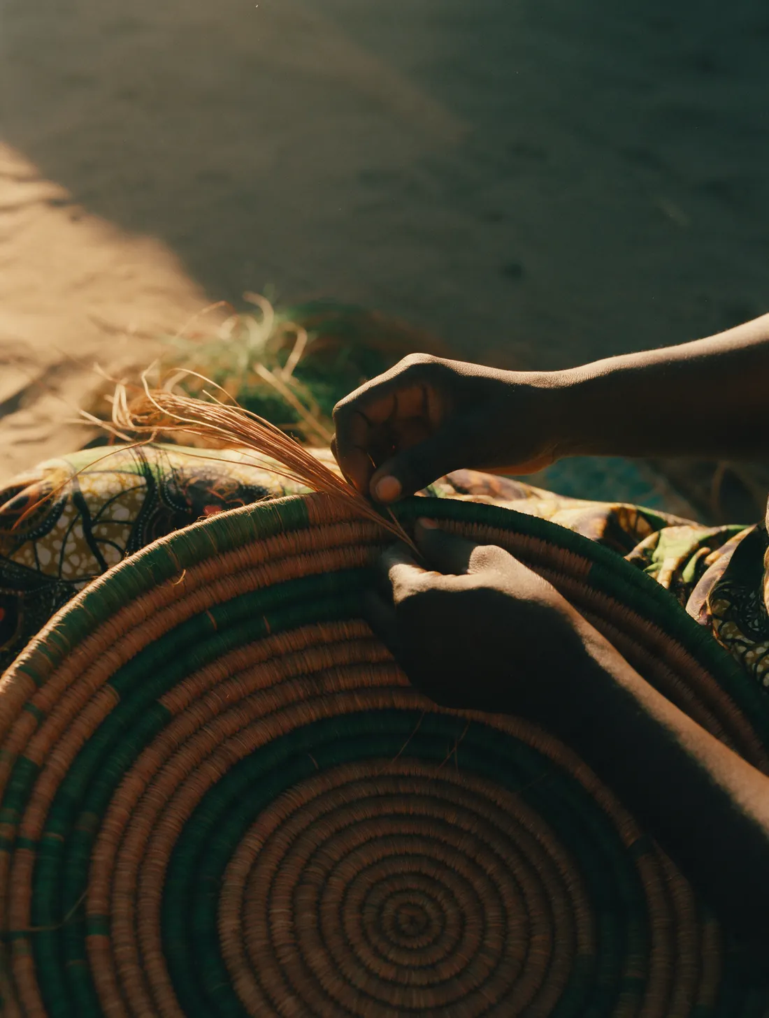 Hands weaving a coiled basket from ochre and green fibres.