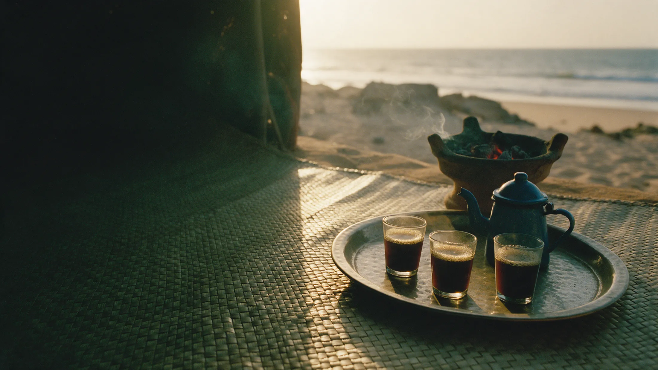 Three small glasses of attaya tea on a metal tray beside a teapot and a charcoal brazier, on a mat by the sea.